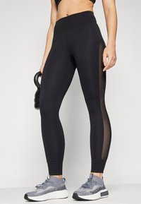 Leggings de sport noirs avec des panneaux en mesh sur les côtés, taille haute et coupe ajustée. Associés à des baskets grises avec un design texturé.