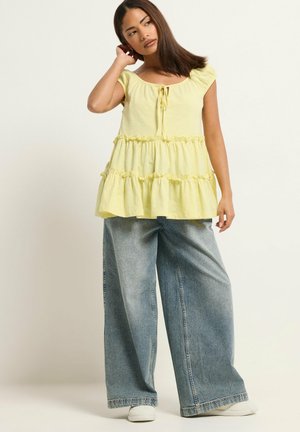 Junge Frau mit langen dunklen Haaren, die ein gelbes, ärmelloses, gerafftes Oberteil und weite blaue Jeans trägt, steht mit einer Hand am Hals.