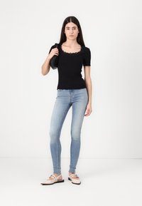Černý přiléhavý top s vlnitým výstřihem, zkombinovaný se světle modrými skinny džíny a béžovými sandály na podpatku. Model drží černou tašku.