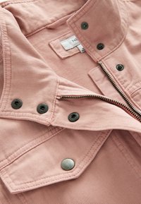 Veste en denim rose avec fermeture éclair partiellement ouverte, boutons-pression en métal et étiquette intérieure visible.