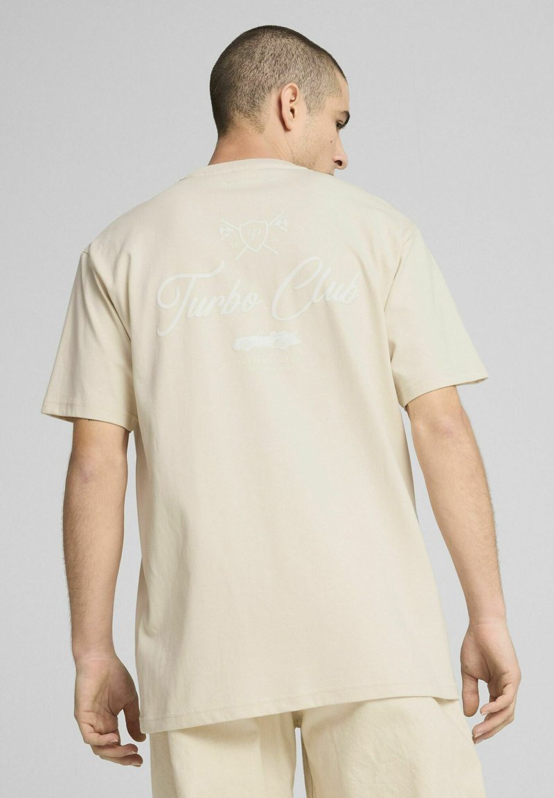 Beige bomulds T-shirt med ryggrafik med teksten "Turbo Club", ledsaget af ikoner og en bilillustration. Korte ærmer, løs pasform.
