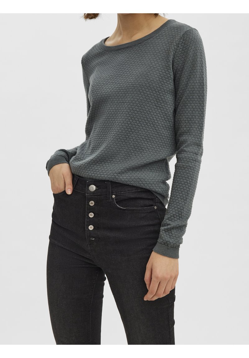 Femme portant un haut gris texturé à manches longues et un jean noir taille haute avec cinq boutons argentés visibles, debout devant un fond uni.