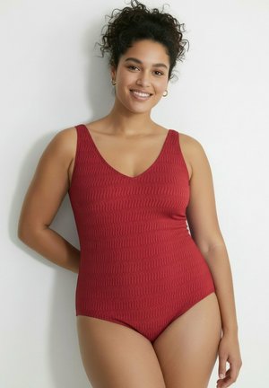 Kobieta o kręconych włosach uśmiechająca się, ubraną w teksturowany czerwony jednoczęściowy strój kąpielowy bez rękawów, stojąca na jednolitym białym tle.