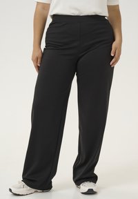 Pantalons larges taille haute noirs avec une texture lisse, présentant un design uni et une taille élastique pour le confort, associés à des baskets blanches.