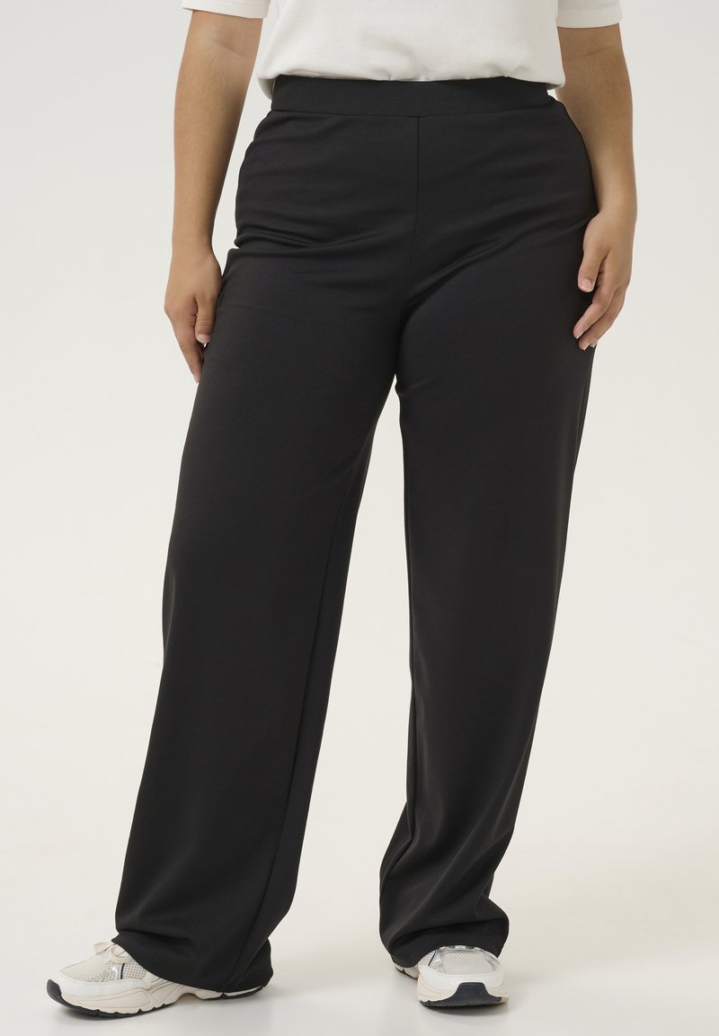 Pantalons larges taille haute noirs avec une texture lisse, présentant un design uni et une taille élastique pour le confort, associés à des baskets blanches.