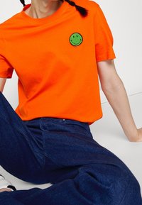 Personne portant un t-shirt orange vif avec un patch visage souriant vert, jean bleu foncé, assise avec un bras étendu en arrière pour le soutien.