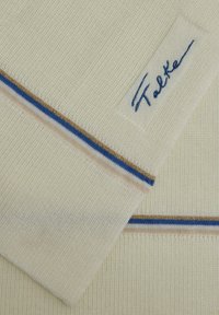 Pull en crème à col rond avec texture côtelée. Présente des rayures bleues et dorées le long de l'ourlet et une étiquette blanche emboutie avec "Falke".