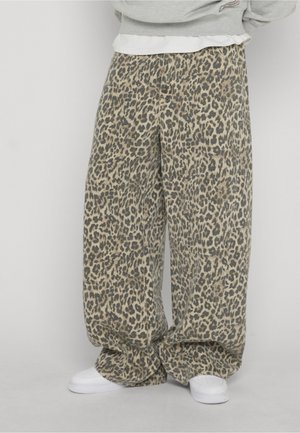 Løse leopardmønstrede bukser iført hvide sneakers og en grå sweatshirt, stående mod en ensfarvet lys baggrund.