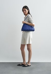 Πλεκτή μπλέ δερμάτινη τσάντα σε σχήμα χόμπο, με μία ενιαία λουρί. Το μοντέλο φοράει μία ριγέ μπλούζα και μπεζ σορτς, στέκεται σε γκρίζο πάτωμα.