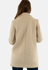 Cappotto in misto lana beige con colletto strutturato, maniche lunghe, texture morbida e taglio dritto, che termina appena sotto i fianchi.