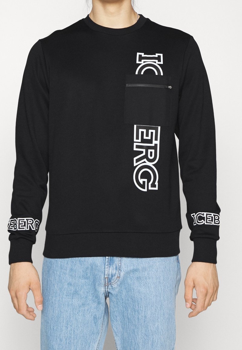 Sweat-shirt noir à manches longues avec texte vertical blanc "ICEBERG" sur le devant et les poignets, avec une poche poitrine zippée, porté avec un jean bleu.