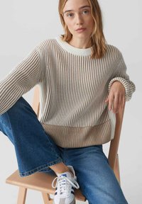 Strickpullover in Creme- und Beigetönen mit gerippten Bündchen, getragen mit blauen Schlagjeans und weißen Sneakers. Sitzt auf einem Holzstuhl.