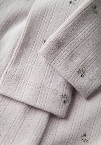 Tissu tricoté beige clair avec une petite broderie de fleurs violettes et des motifs texturés côtelés, montrant des manches et des poignets repliés.