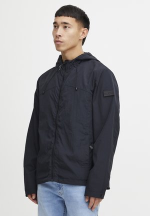 Veste légère noire avec capuche, fermeture éclair et cordon de serrage. Comprend un tissu texturé, des poches avant et un patch de marque sur la manche.