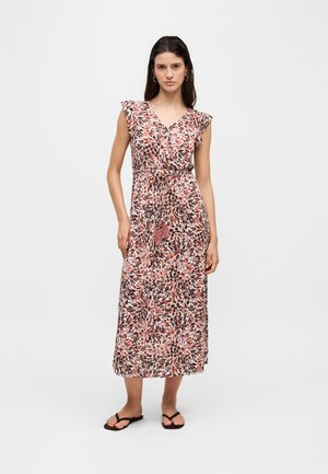 Kvinde stående iført en ærmeløs blomster maxi-kjole med flæsedetaljer på skuldrene og frynsebælte, kombineret med sorte flip-flop sandaler.