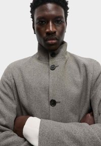 Abrigo de lana gris con un cuello alto, que cuenta con botones negros y un corte holgado. La chaqueta tiene un acabado texturizado y mangas anchas.