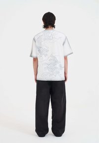 Oversized biały t-shirt z krótkim rękawem z szarym nadrukiem smoka na plecach, w zestawieniu z luźnymi czarnymi dżinsami, tworzącymi swobodną sylwetkę.