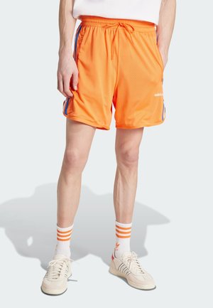 Pantalones cortos deportivos de malla naranja con cintura elástica, rayas laterales en azul y un logotipo en el lado inferior. Llevados con calcetines blancos y zapatillas.