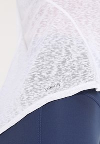 Haut blanc, léger et semi-transparent avec un motif texturé et une étiquette "CLIMALITE" à l'ourlet, associé à un legging de sport bleu foncé.
