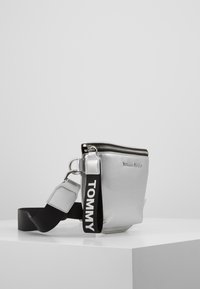 Borsa a tracolla sintetica argento con chiusura a zip, tracolla nera regolabile, hardware in metallo e dettaglio del logo impresso sulla parte anteriore.