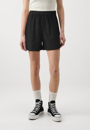 Personne portant un short noir à cordon, des chaussettes blanches et des baskets montantes noires, debout devant un fond uni.