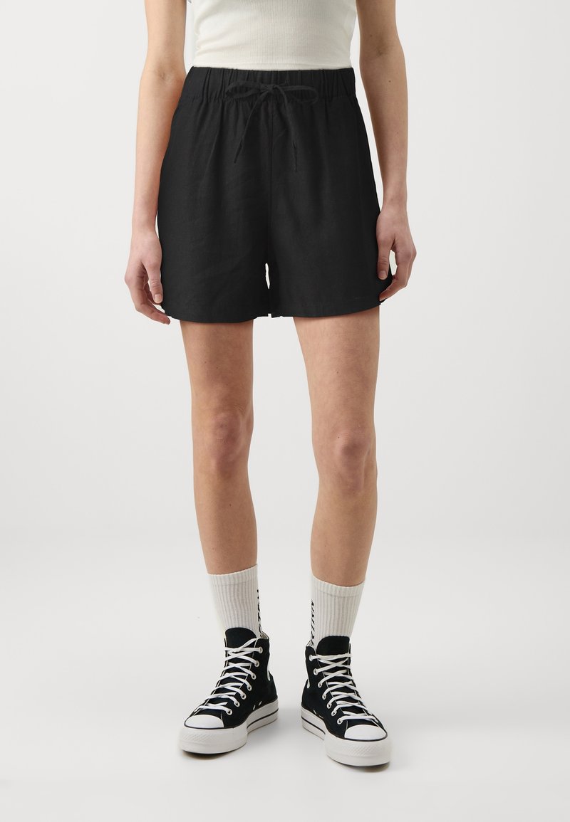 Personne portant un short noir à cordon, des chaussettes blanches et des baskets montantes noires, debout devant un fond uni.