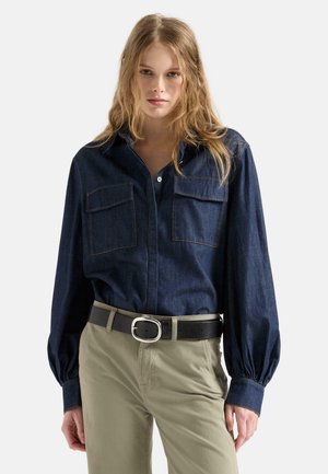 Woman wearing a dark denim shirt with pockets and beige pants, standing against a plain light background.