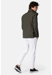 Oliwkowa kurtka z wysokim kołnierzem, bocznymi kieszeniami i zapinanymi mankietami, w połączeniu z białymi spodniami i czarnymi trampkami. Gładki, trwały materiał.