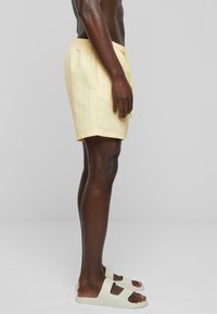 Shorts de bain jaune clair avec de fines rayures verticales, dotés d'une taille élastique et de poches latérales, associés à des sandales à enfiler de couleur pâle.