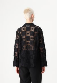 Svart spetsblus med ett öppet mönster med fyrkantiga motiv, knappade manschetter och krage. Baksidan är halvgenomskinlig.