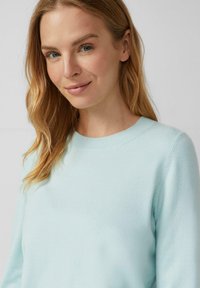 Młoda kobieta z blond włosami i niebieskimi oczami, ubrana w jasnoniebieski sweter z okrągłym dekoltem, lekko uśmiechająca się na tle jednolitego koloru.