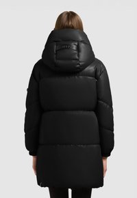 Persoon gekleed in een zwarte, gewatteerde lange donsjas met een grote capuchon, gezien van achteren tegen een effen achtergrond.