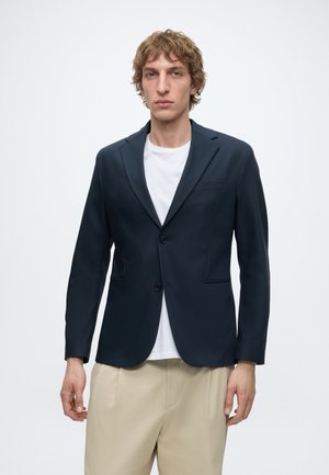 Ung mand med krøllet hår iført en mørk marineblå blazer over en hvid skjorte og beige bukser, stående mod en ensartet lys baggrund.