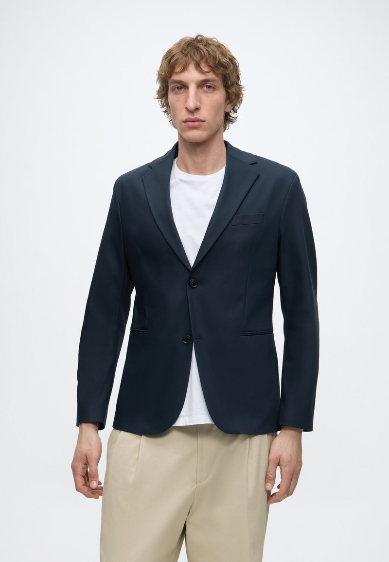 Jeune homme aux cheveux bouclés portant un blazer bleu marine foncé par-dessus une chemise blanche et un pantalon beige, debout sur un fond clair uni.