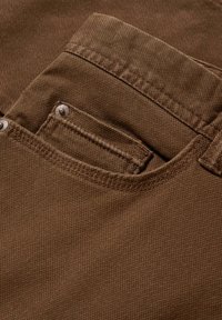 Gros plan sur la poche d'un jean en denim marron avec une couture visible et des rivets métalliques sur une texture de tissu grossier.