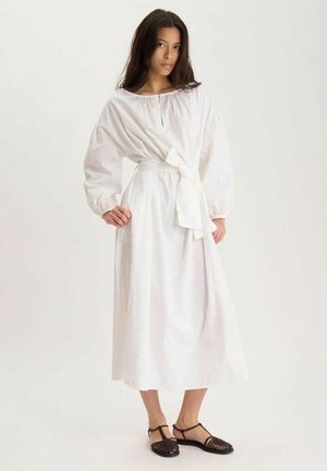 Jeune femme portant une longue robe blanche avec des manches bouffantes et une taille nouée, debout avec une main sur la hanche, sandales plates noires.