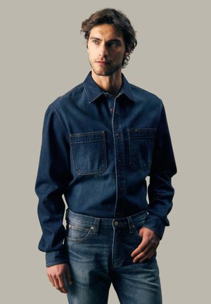 Mann mit kurzen lockigen Haaren, der ein dunkelblaues Jeanshemd und Jeans trägt und vor einem einfarbigen beigen Hintergrund steht.