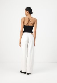 Schwarzes ärmelloses Oberteil mit gekreuzten Rückenträgern, kombiniert mit locker sitzenden weißen Hosen. Model trägt schwarze flache Schuhe.