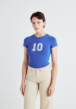 Royal blue short-sleeve bodysuit with a white number "10" printed on the front, paired with light beige high-waisted jeans.