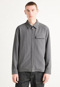 Chaqueta gris con cremallera, de tejido texturizado, cuello con solapa, un bolsillo en el pecho y manga larga. Combinada con pantalones de mezclilla oscuros.