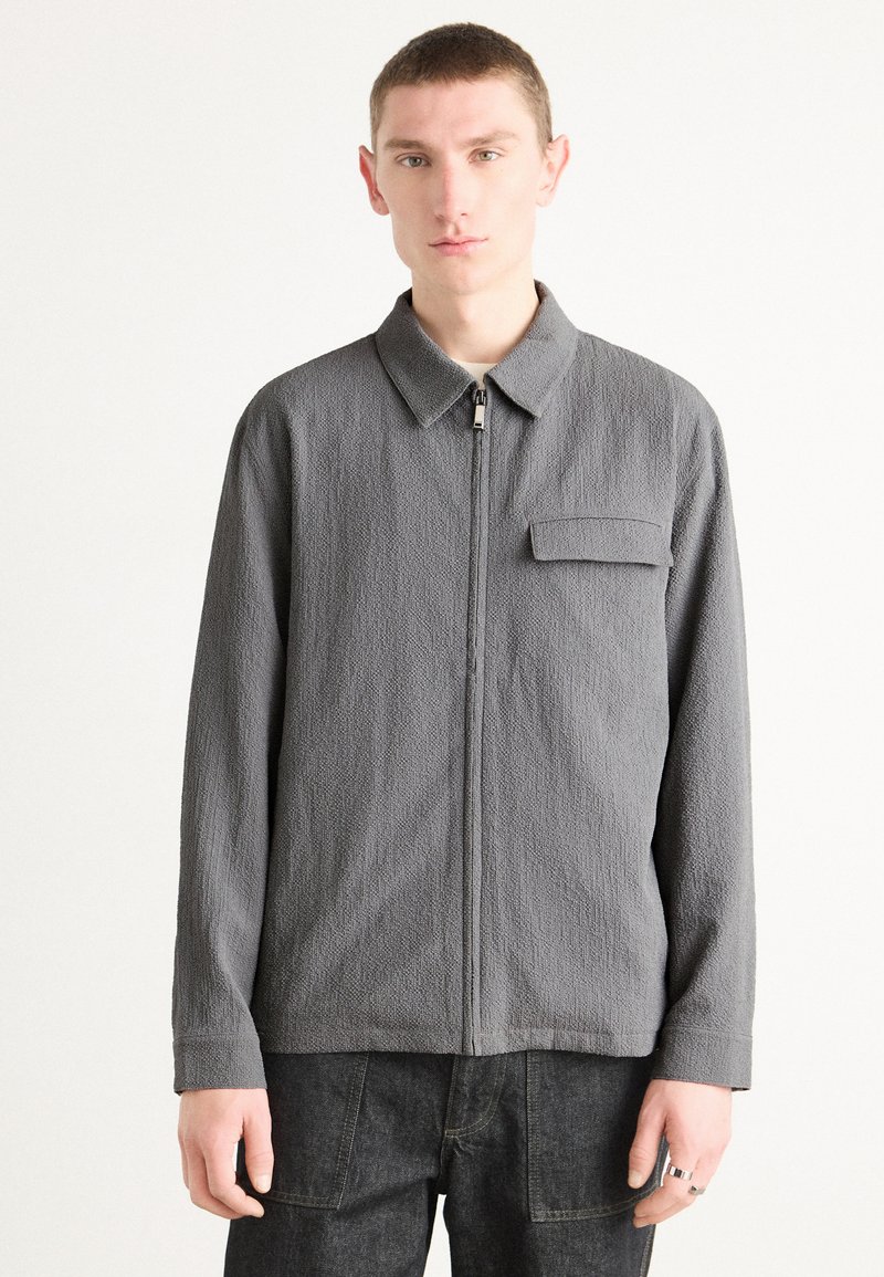 Chaqueta gris con cremallera, de tejido texturizado, cuello con solapa, un bolsillo en el pecho y manga larga. Combinada con pantalones de mezclilla oscuros.