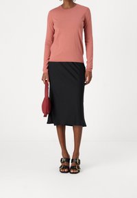 Langärmliges korallenfarbenes Oberteil kombiniert mit einem schwarzen, eng anliegenden Rock, accessorized mit einer roten Tasche und schwarzen Sandalen mit goldenen Schnallen.