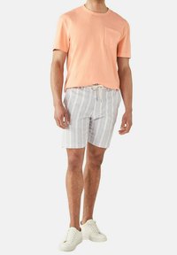 Maglietta in cotone pesca con tasca, abbinata a pantaloni corti a righe blu chiaro. Le scarpe sono bianche con design a lacci. Look semplice e informale.