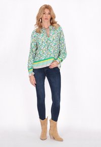 Blusa verde con motivo floreale e scollatura a chiave, polsini con bordo in pizzo, abbinata a jeans blu scuri e stivali da cowboy beige con tacco.