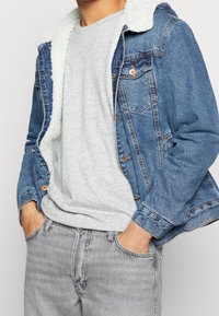 Jeansjacke mit Kunstfellfutter, blaue Farbe, mit zwei Brusttaschen und Kupferbeschlägen. Getragen über einem grauen T-Shirt und grauen Jeans.