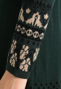 Grön stickad tröjärm med invecklade beige och marinblå mönster, inklusive blommotiv och geometriska mönster. Mjuk textur, mudd med ribbad kant.