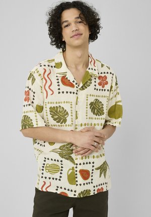 Jeune homme aux cheveux bouclés portant une chemise beige à manches courtes avec des motifs de feuilles et de fruits tropicaux verts et rouges, les mains jointes à la taille.