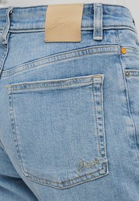 Φόρμα τζιν σε ανοιχτό μπλε χρώμα με δερμάτινο σημείο στη μέση, που φέρει κεντημένο λογότυπο, πίσω τσέπες και κίτρινα διακοσμητικά ραφές αντίθετου χρώματος.