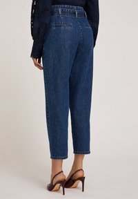 Jeans de mezclilla azul de tiro alto con pierna recta, bolsillos traseros y costuras visibles. Combinados con zapatos de tacón burdeos.