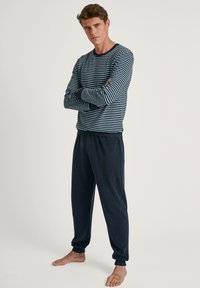 Gestreepte marineblauwe en lichtblauwe langverwarmde shirt met ronde hals; gecombineerd met donker marineblauwe pyjamabroek. Zachte stof, losse pasvorm, elastische manchetten.